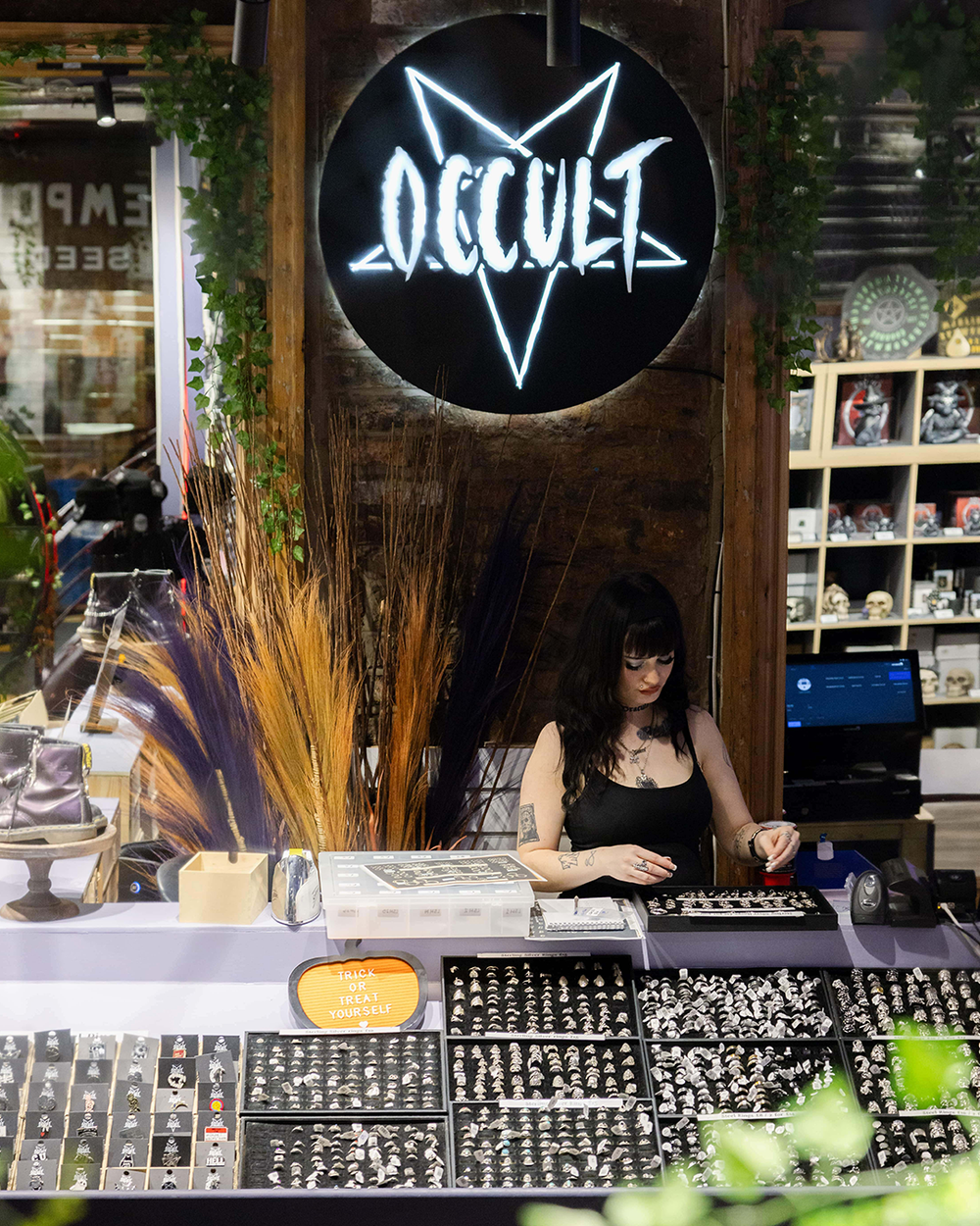 A woman in a black top stands behind a counter at a gothic-themed shop, with a large illuminated 'Occult' sign above her. The counter displays many small jewelry pieces, and shelves with figurines are visible in the background.